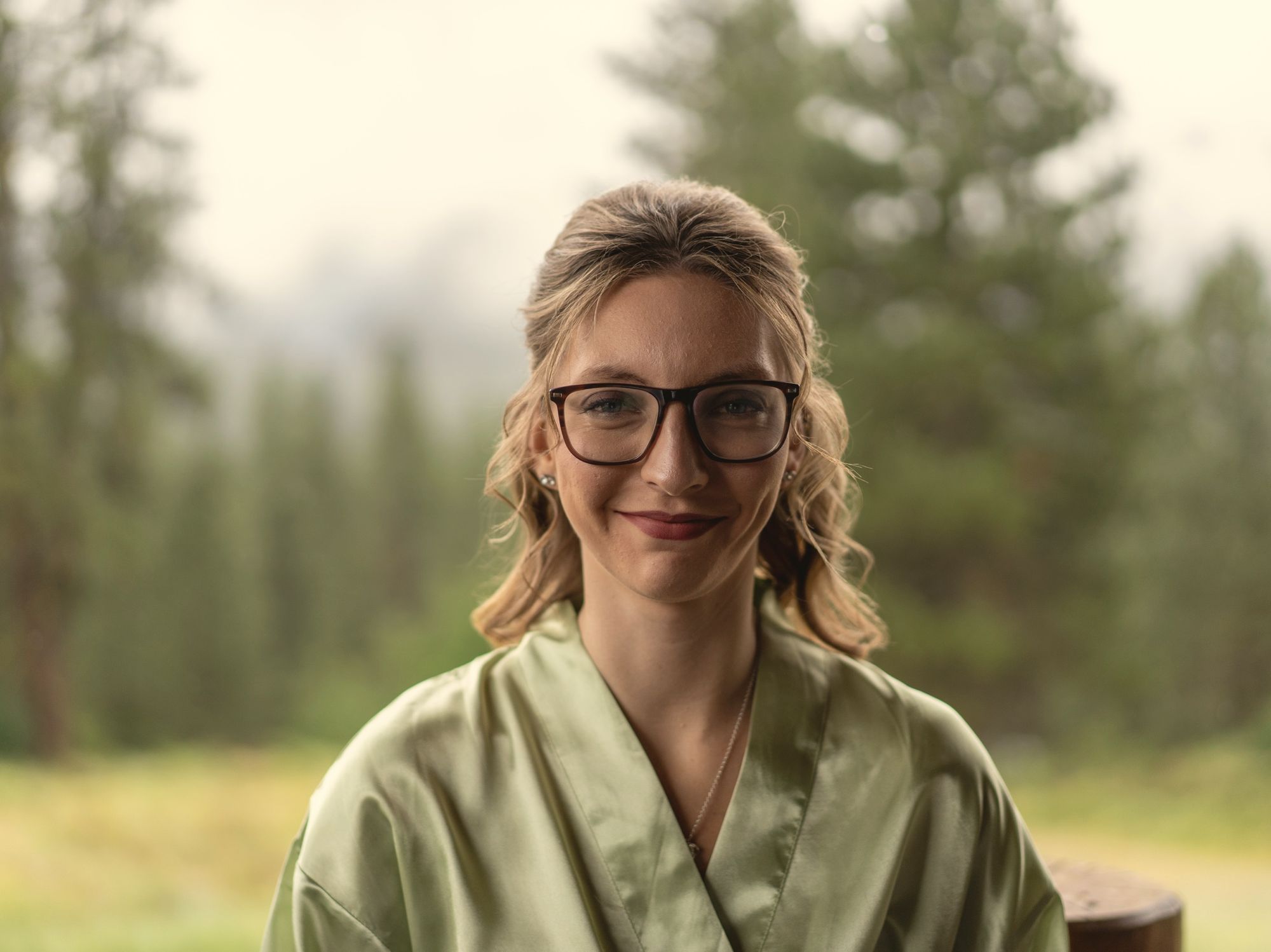 Stephanie smiling softly in a sage-green top against misty pine trees